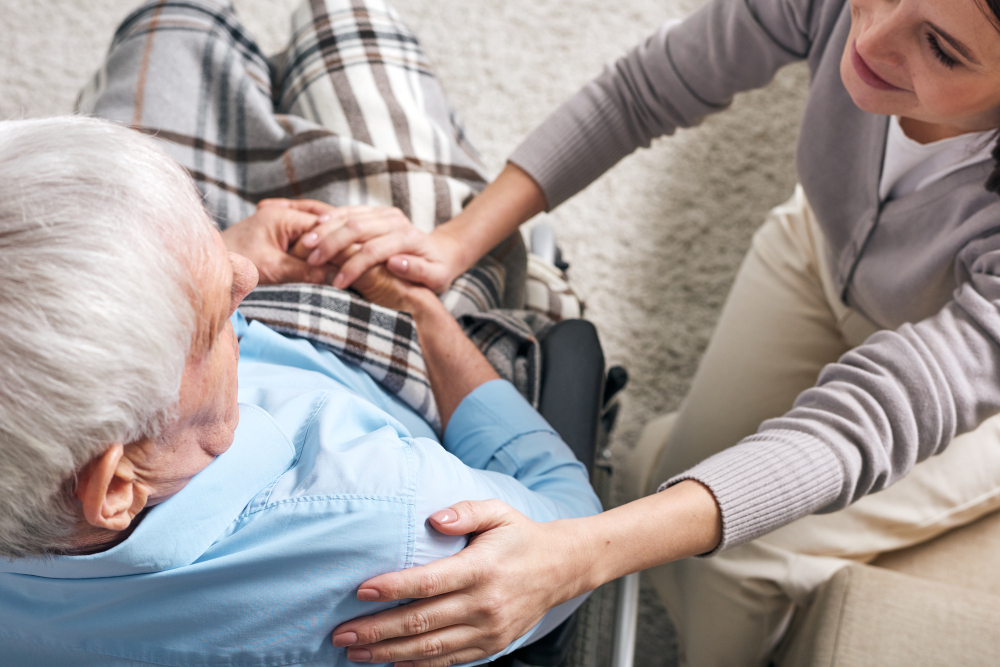 young-supportive-female-caregiver-sitting-by-senior-man-wheelchair-keeping-her-hand-his-shoulder-while-comforting-him